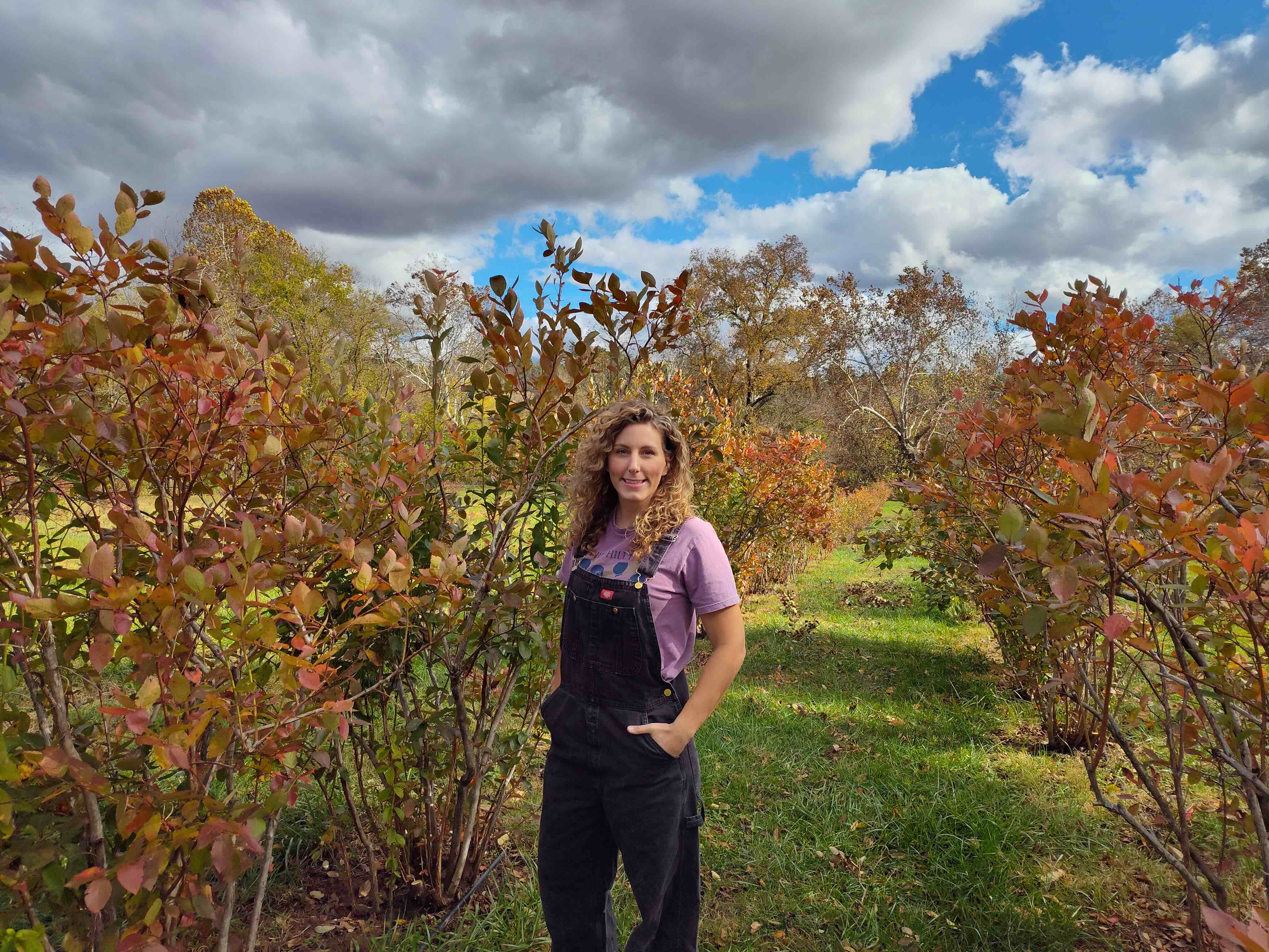 Young woman in overalls stands next to blueberry bushes