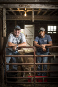Two farmers stand smiling with their elbows resting on a barn gate. They stand inside a barn. A goat is sniffing one of their hands. 