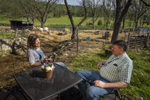 A shepherd and a service provider sit at a table outside. Sheep graze in the background.